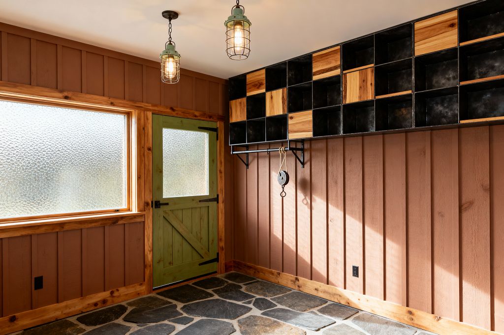 A rustic mudroom features terracotta board-and-batten walls with honey knotty-pine trim, a sage green barn-style door with a frosted glass inset, and a large privacy-glass window. The floor is irregular charcoal slate flagstone; two cage-pendant lights hang from a white ceiling, and a wall of blackened-steel cubbies with natural wood panels and a mounted rail with rope-and-pulley hook offer storage and a handmade, industrial touch.