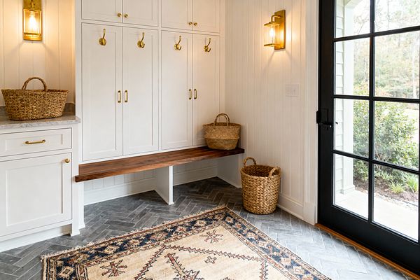 A bright transitional mudroom features white Shaker cabinets with brass pulls and hooks, a thick walnut bench, and vertical beadboard walls flanked by two brass-and-glass sconces. A marble-topped base cabinet holds a woven basket, while charcoal gray herringbone tile is layered with a tan and navy vintage-style rug, and a black grid-paneled glass door opens to the garden with additional woven baskets providing storage below.