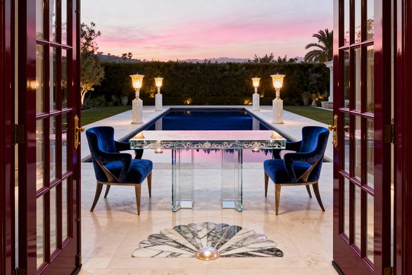 Through burgundy French doors, a Hollywood Regency patio frames a rectangular navy pool, flanked by four glowing urn lanterns on fluted marble pedestals. In the foreground, a mirrored console with etched floral motifs stands between two royal blue velvet armchairs on cream limestone, set over a fan-shaped black-and-white marble inlay, with clipped hedges, palm silhouettes, and a pink sunset reflecting in the water.