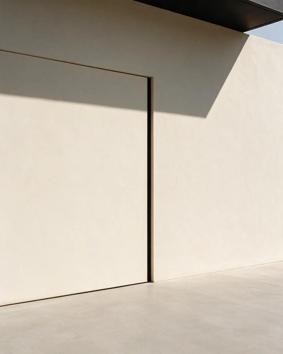 A minimalist exterior featuring smooth cream stucco walls, a large flush door or panel outlined by thin dark shadow gaps, and a pale concrete floor. Strong sunlight casts sharp geometric shadows from a dark roof overhang above, creating a calm, understated architectural composition.