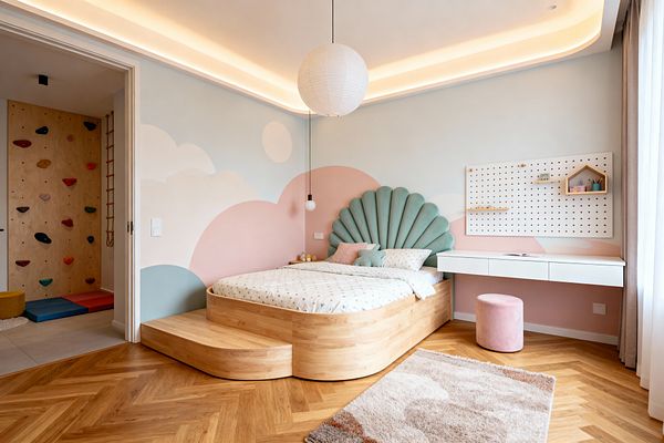 Contemporary kids’ bedroom featuring a rounded light-oak platform bed with an integrated step and a seafoam scalloped velvet headboard against a pastel cloud mural in blush, soft blue, and cream. The room has herringbone oak flooring, a plush beige rug, a floating white desk with pegboard, a pink velvet stool, paper lantern and globe pendants, sheer curtains, and an adjacent play nook with a birch plywood climbing wall, rope ladder, and colorful mats.