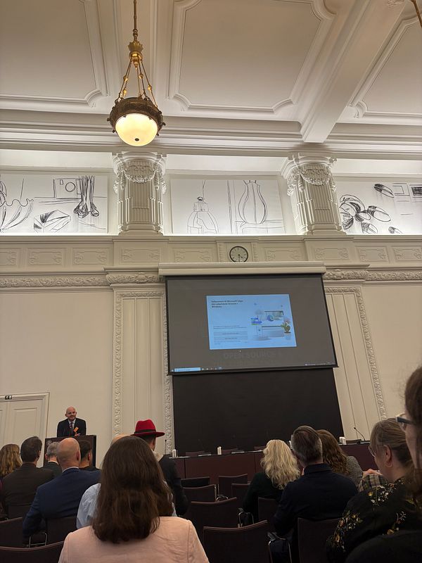 A speaker presents at a conference with a large screen displaying a presentation in an ornate room.