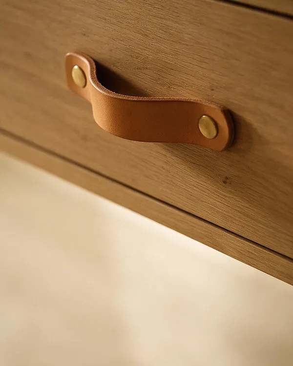A close-up of a warm wood drawer front with visible grain, fitted with a tan leather pull secured by two brass rivets. The lower portion fades into a soft cream background, emphasizing the natural materials and minimalist Organic Modern aesthetic.
