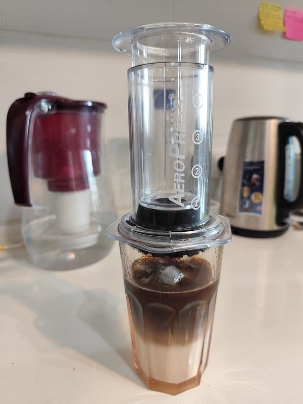An AeroPress coffee maker is positioned above a glass filled with layered coffee and milk.