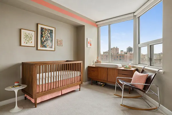 A bright nursery with a warm wood crib against a soft beige wall, framed artwork above it, and a long wooden dresser beneath large corner windows. A chrome and leather rocking chair sits by the window, while a small side table and pink patterned crib skirt add playful contrast.