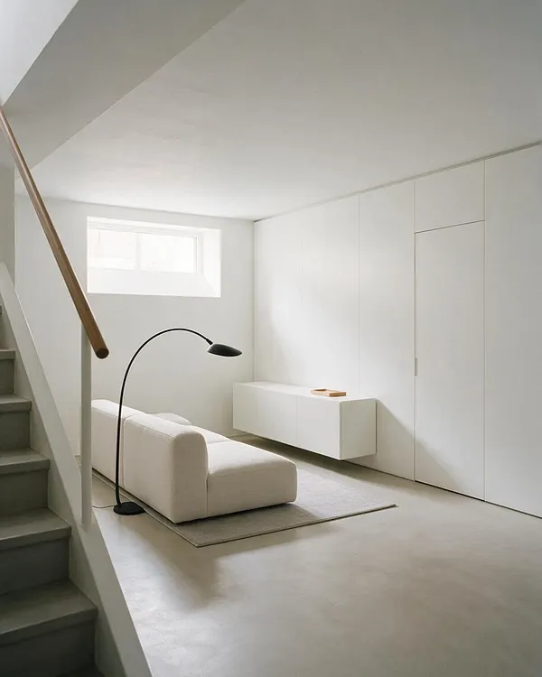 A bright minimalist basement lounge with white walls and a low white sofa on a small rug. A black arched floor lamp stands beside the seating, while a long wall of seamless built-in cabinets and a small horizontal window keep the space clean and uncluttered. 