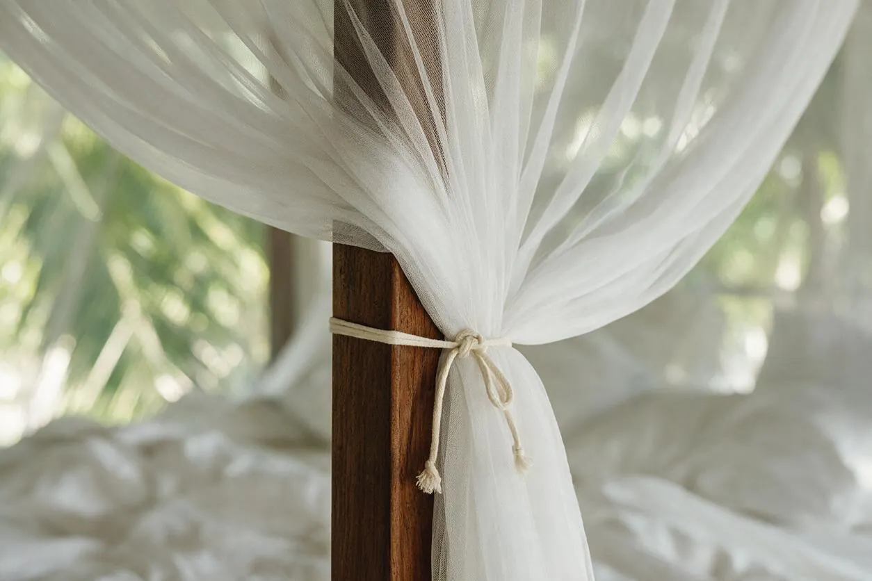 A close-up of a tropical bed canopy with airy white sheer fabric gathered around a dark wooden post and tied with a thin cord. Soft bedding and blurred palm greenery in the background create a relaxed, breezy resort feel.