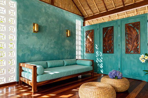 Sunlit tropical media lounge with a long teak daybed upholstered in seafoam fabric and bolster cushions, placed against textured turquoise walls. Carved wooden leaf panels inset in matching turquoise screens face two woven rattan ottomans on a dark wood floor. White breeze-block walls filter daylight, casting patterns across the room, while brass sconces and a palm-thatch ceiling add warmth; a vase of plumeria and a hydrangea bouquet provide fresh color.