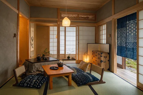 A calm Japanese Zen entryway with tatami mat flooring, low wooden table, floor cushions, shoji screens, and warm paper lantern lighting. Natural wood framing, a blue noren curtain, and a small chest create a peaceful, minimalist atmosphere.