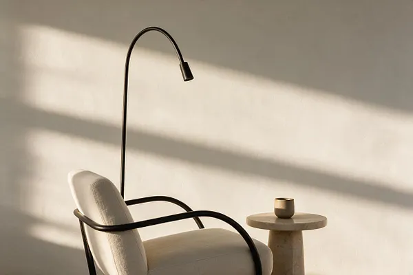 A minimalist corner features a cream upholstered lounge chair with slim black metal arms beside a small round pedestal table in pale stone. An arched black floor lamp rises behind the chair, while warm sunlight casts soft horizontal shadows across the off-white wall.