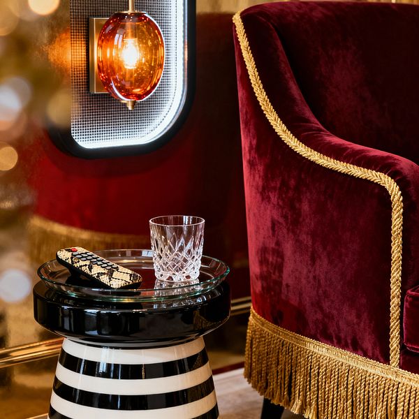 A Hollywood Regency media nook features a crimson velvet armchair trimmed with gold braid and bullion fringe beside a black-and-white striped pedestal table with a clear glass top. A cut-crystal tumbler and snakeskin-pattern remote sit on the table. On the wall, an amber glass globe sconce with a brass mount and backlit mesh panel casts warm light across glossy gold walls, creating a glamorous, intimate ambiance for film watching.