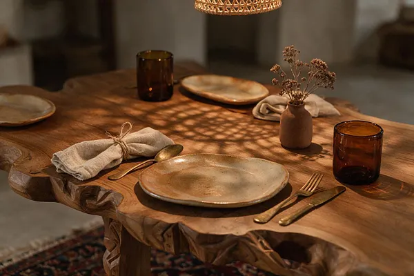 A warmly lit dining table features a live-edge wooden top with carved edges, set with cream ceramic plates, brass cutlery, folded linen napkins, amber glass tumblers, and a small clay vase with dried flowers. Patterned shadows from a woven pendant lamp fall across the surface, creating a cozy bohemian atmosphere.