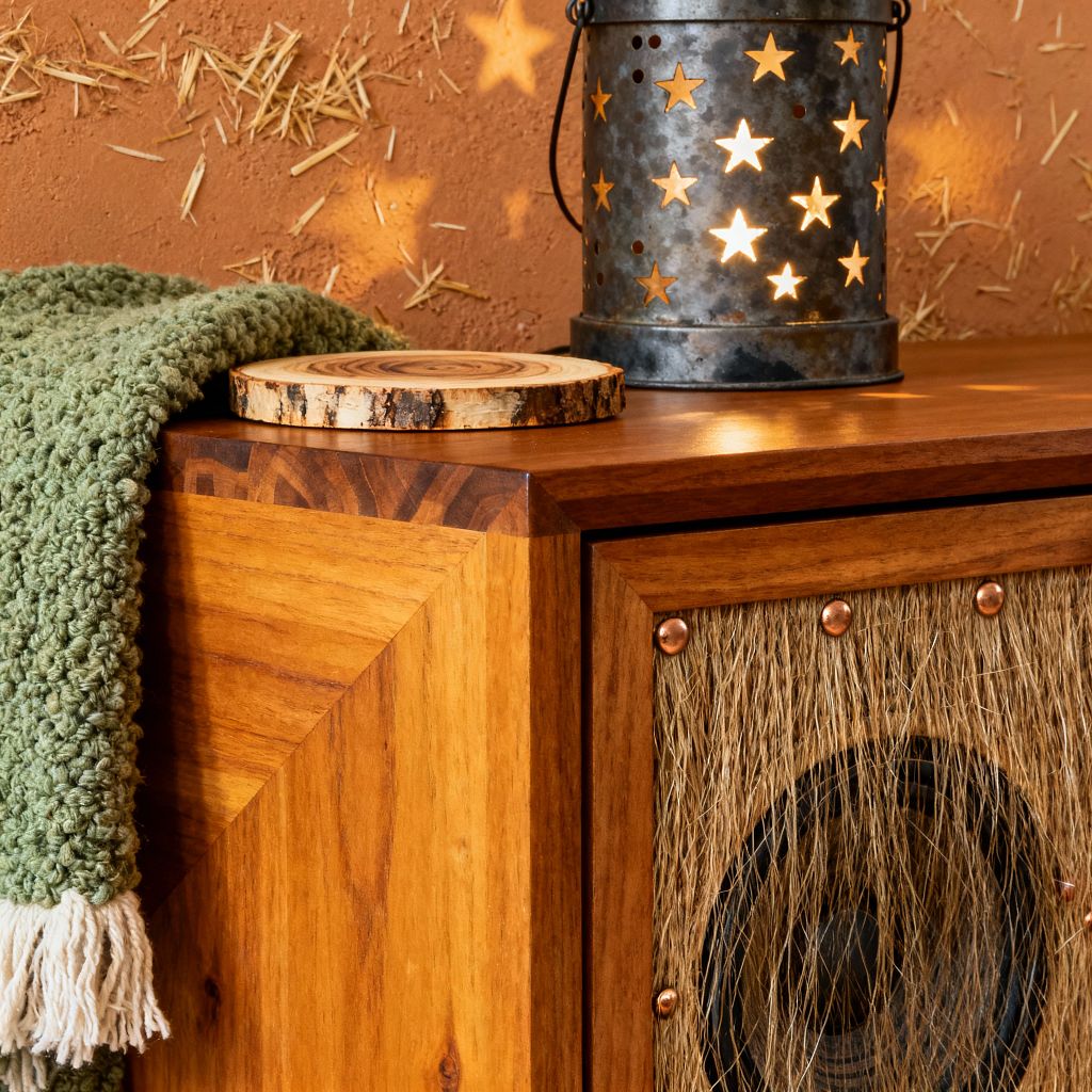 Close-up of a rustic media console made of honey-toned wood with mitered corners. A speaker sits behind a jute fiber grille secured with copper rivets. On top, a chunky sage green knit throw with cream fringe lies beside a live-edge wood slice. A galvanized metal lantern with star cutouts casts warm light and star-shaped shadows across a terracotta, straw-flecked plaster wall, creating a cozy, handcrafted atmosphere.