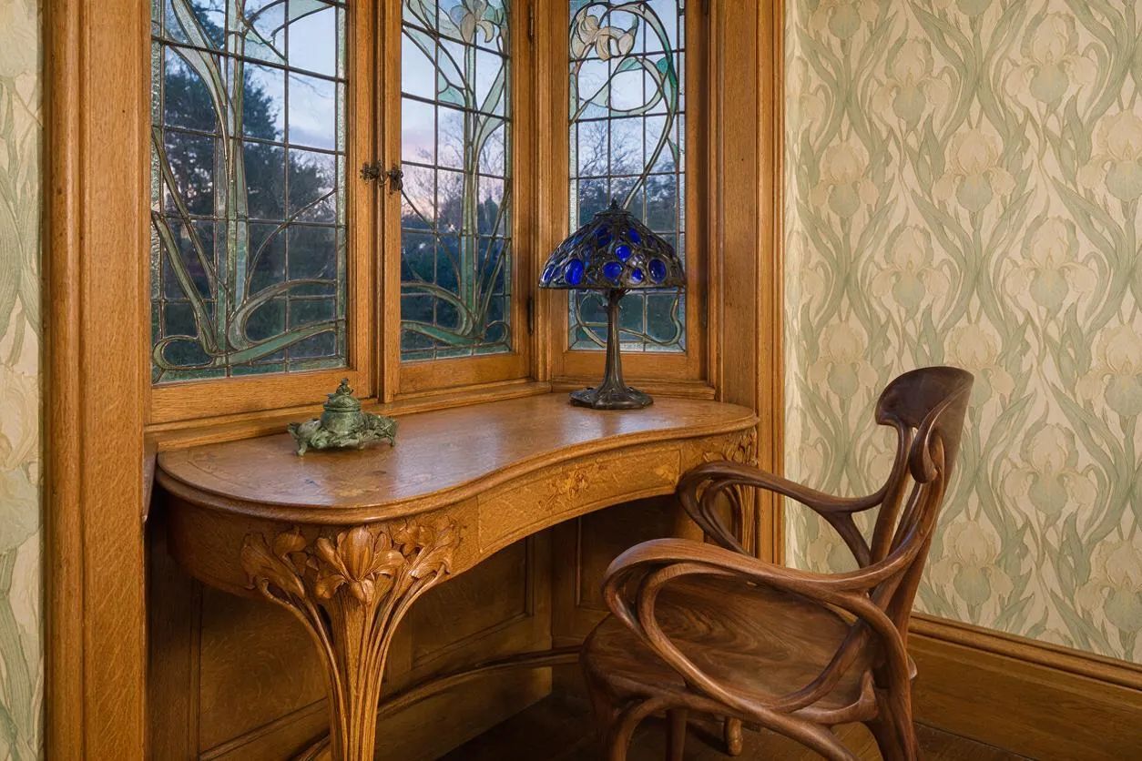 A sunroom corner in Art Nouveau style features a built-in carved wood desk beneath leaded glass windows with flowing floral motifs. A sculptural wooden armchair sits beside a blue glass lamp, with patterned botanical wallpaper and warm wood trim creating a refined, vintage atmosphere.
