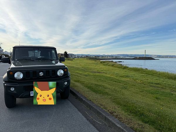 A black Suzuki Jimny parked by a scenic waterfront with a Pikachu graphic on the front.
