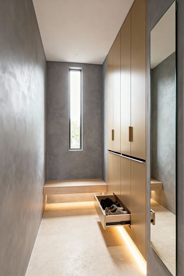 A narrow minimalist mudroom with smooth gray plaster walls and a vertical slit window bringing daylight. On the right, full-height matte sand cabinets with brass pulls and a black finger rail include a pull-out shoe drawer. A pale wood floating bench wraps the back wall, while warm LED strip lighting runs under the bench and cabinets above a light limestone floor, opposite a full-height mirror.