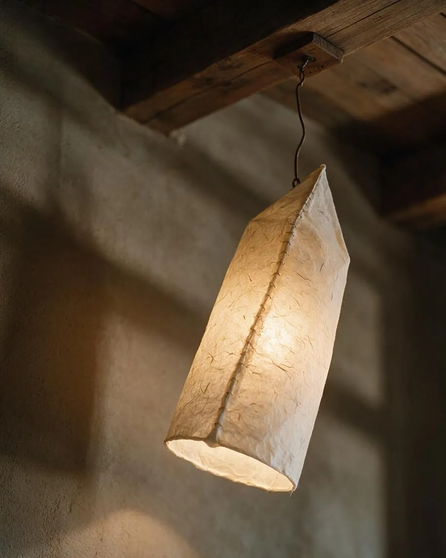 A hanging pendant light made from textured handmade paper is suspended by a thin dark cord from a wooden beam. The soft warm glow reveals the paper’s natural fibers and stitched seam against a muted plaster wall.