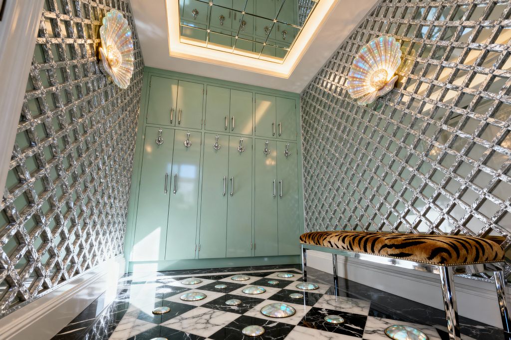 A glamorous mudroom in Hollywood Regency style showcases glossy mint built-in lockers with polished chrome hooks and long pulls, flanked by diamond-pattern mirrored wall panels. The space features shell-shaped iridescent sconces, a mirrored coffer ceiling with warm cove lighting, a black-and-white marble checkerboard floor dotted with circular mother-of-pearl inlays, and a chrome-legged bench upholstered in tiger-print velvet.