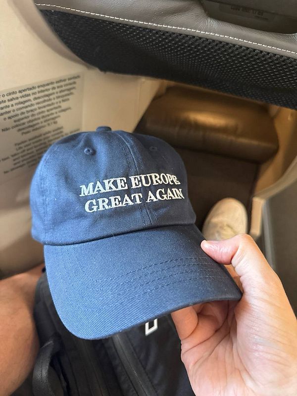 A hand holds a blue cap with the phrase 'MAKE EUROPE GREAT AGAIN' prominently displayed.