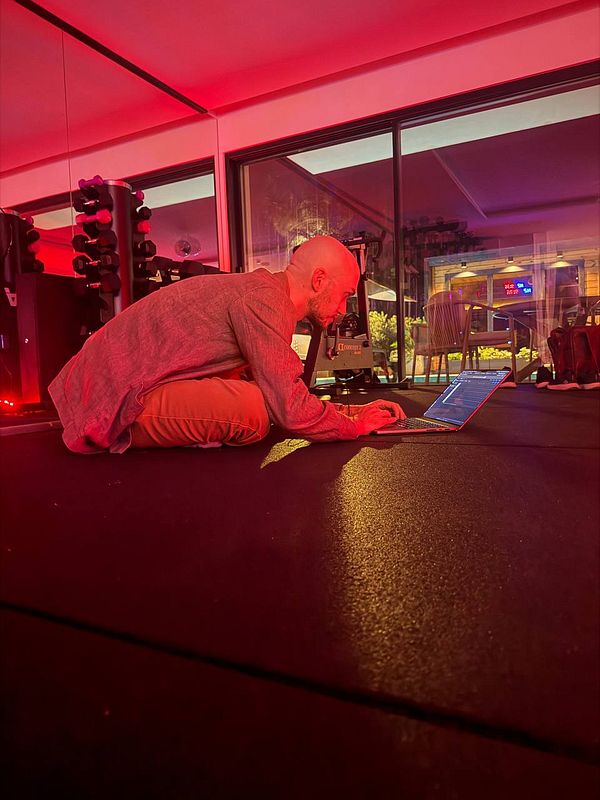 A man kneeling on a gym floor working on a laptop with red ambient lighting around him.