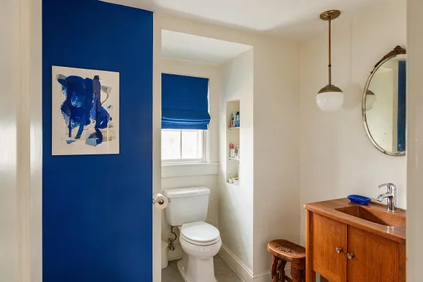 A compact powder room with a vivid cobalt-blue accent wall, abstract blue artwork, and a white toilet tucked into a narrow alcove with a blue Roman shade. On the right, a warm wood vanity with a copper-toned sink basin, chrome faucet, round mirror, and brass-and-glass pendant create a mix of modern and vintage details.