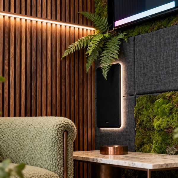 A media room corner features a vertical walnut slat wall with a linear LED strip, adjacent to a charcoal grid of acoustic panels set with bright green moss and fern fronds. A backlit black speaker is mounted below a TV, while a sage green bouclé sofa and a light marble-topped table with a bronze base and copper tray sit in the foreground, creating a warm, nature-inspired space with soft textures and integrated lighting.
