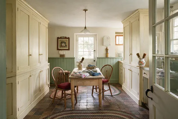 A cottagecore mudroom with tall cream built-in cabinets on both sides, mint green wainscoting, and a small rustic wooden table in the center with folded textiles. Two bentwood chairs sit on patterned rugs over a tiled floor, while a glass-paned door and pendant light add a soft, vintage feel.