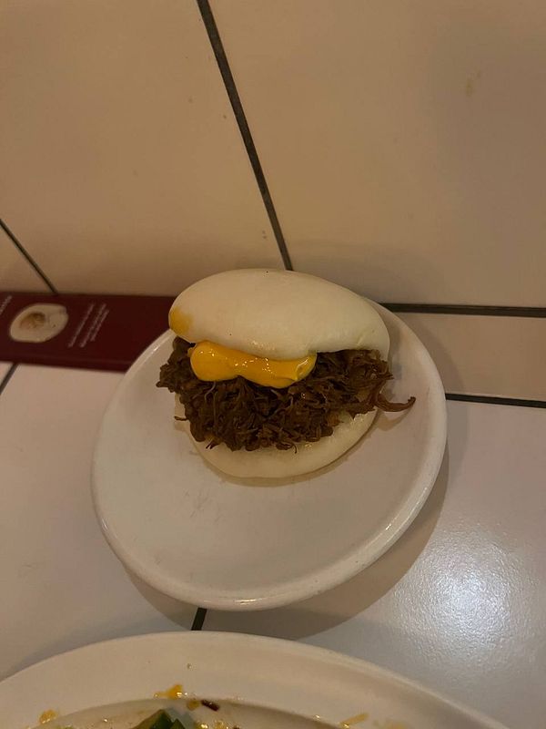 Close-up of a freshly prepared bao bun filled with meat, sauce, and garnished with sesame seeds and greens.