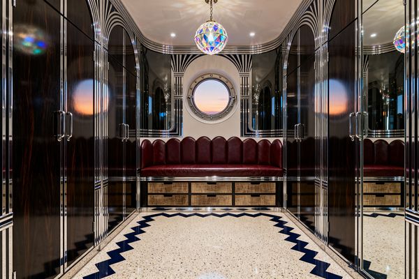 A symmetrical Art Deco mudroom lined with high-gloss dark wood wardrobes trimmed in chrome and mirrored arches. At the end, a built-in bench with a scalloped burgundy leather back sits above cane-front drawers, centered beneath a round porthole window. A terrazzo floor with a navy zigzag border leads to the seat, while an iridescent globe pendant and recessed lights reflect across the lacquered surfaces.