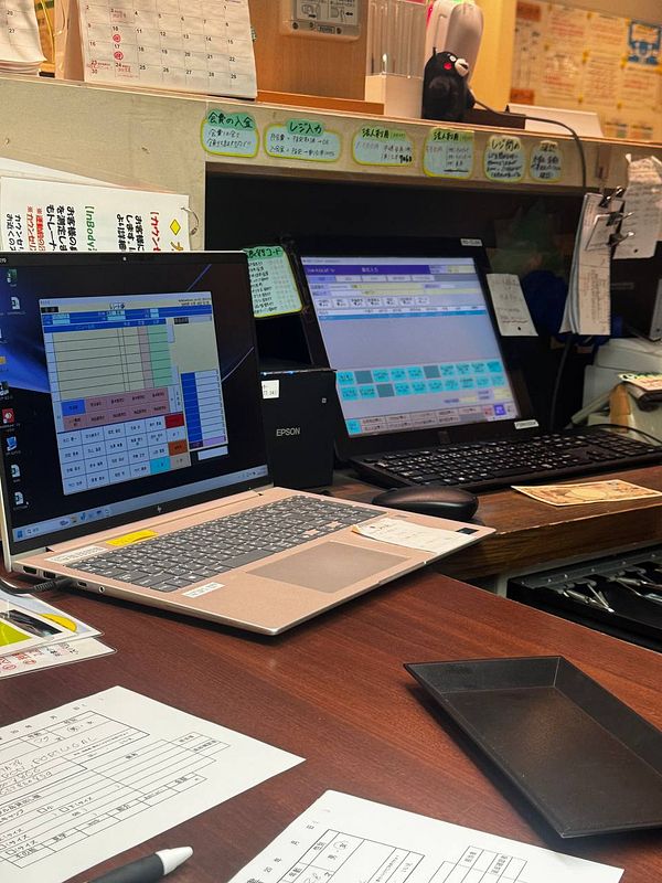 The image depicts a workspace with two laptops, paperwork, and various office supplies in a gym setting in Kyoto, Japan.