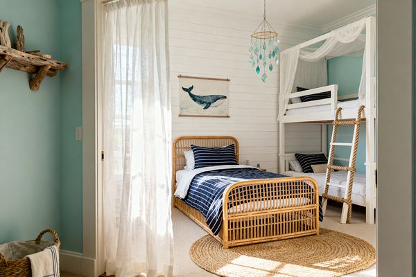 Sunlit coastal kids' room with a natural rattan twin bed in front of a white shiplap wall, navy-and-white striped bedding, and a whale print above. A white bunk bed with draped sheer canopy and a rope-sided ladder stands to the right, while sea-glass pendant, jute rug, driftwood shelf, aqua walls, linen curtains, and baskets complete the beachy look.