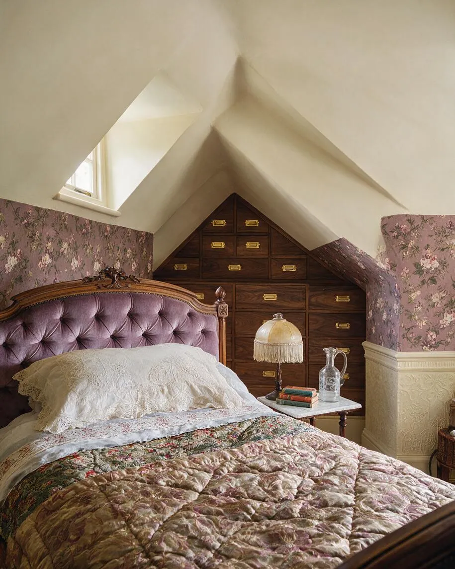 A cozy Victorian-style guest bedroom under a steep pitched ceiling, featuring a carved wooden bed with a mauve tufted velvet headboard and lace pillows. Floral wallpaper covers the lower walls, while a built-in wall of dark wood drawers with brass pulls fills the back gable. A small bedside table holds a fringed lamp and glass pitcher, adding a nostalgic, intimate feel.