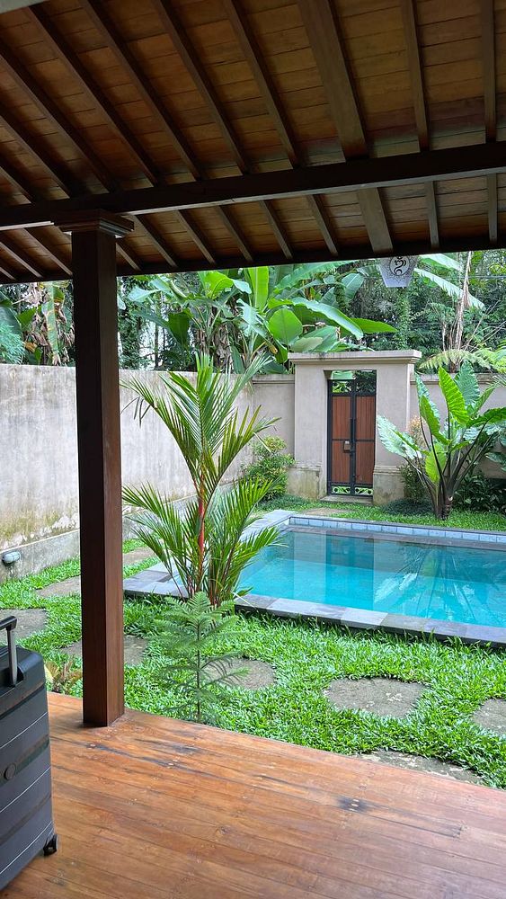A view from a wooden porch overlooking a swimming pool and lush greenery in a villa.