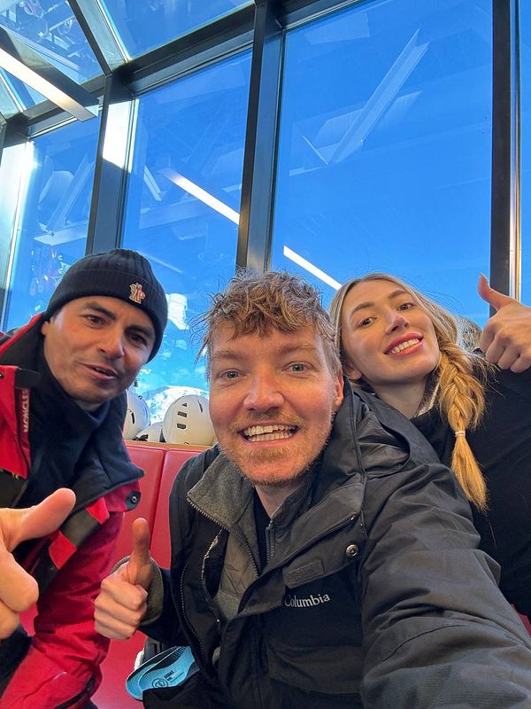 Three friends are taking a selfie in a ski lift with a clear blue sky in the background.