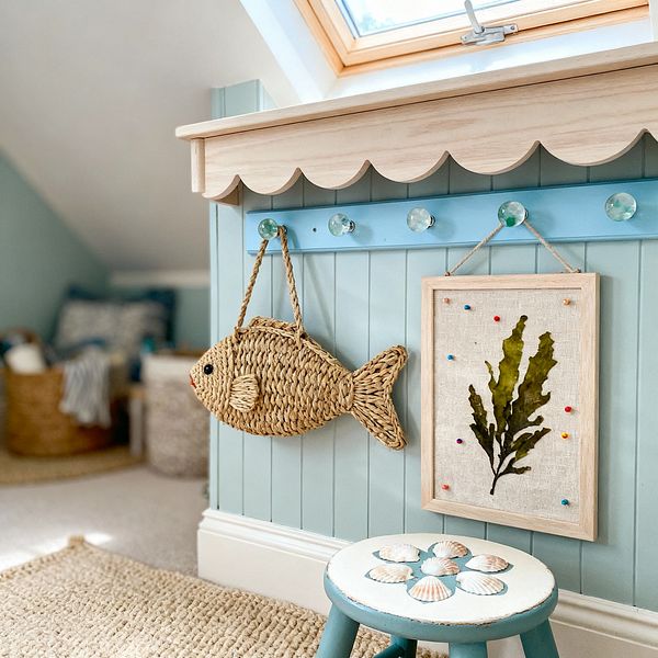 A cozy kids' coastal nook shows seafoam beadboard topped by a scalloped light-wood shelf and a sky-blue peg rail with glass knobs. A woven seagrass fish and a framed linen seaweed print hang above a chunky jute rug; a teal painted stool with shell motifs sits nearby, while a sloped ceiling and wood-trimmed skylight bring natural light to rattan baskets and soft, beachy textiles.