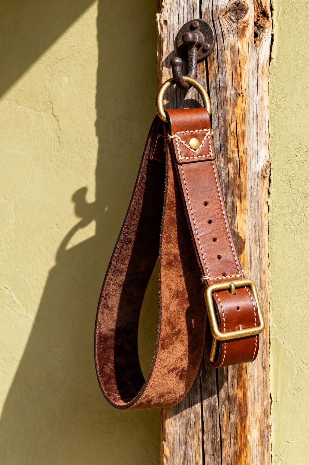A close-up of a brown leather weightlifting belt with cream stitching and a brass buckle hanging from a blackened iron double hook mounted on a weathered reclaimed wood post. A sunlit, textured sage-green plaster wall forms the backdrop, casting a crisp shadow of the hook and strap. The scene conveys rustic home gym storage with durable, natural materials and visible patina that blends warmth, utility, and handcrafted detail.