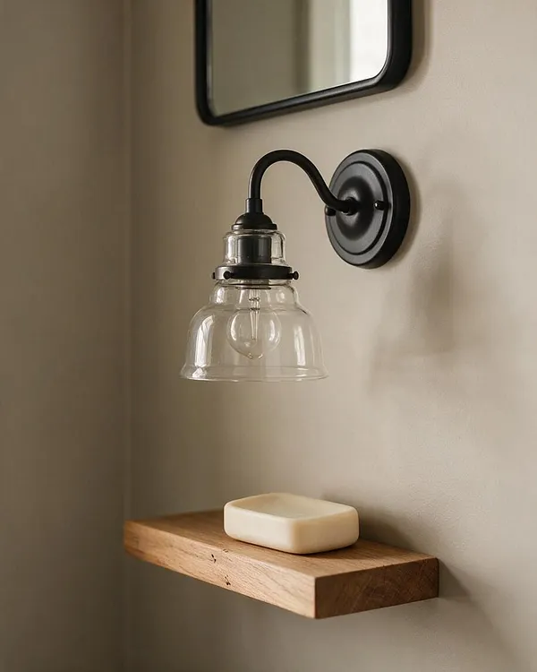 A close-up of a farmhouse-style wall sconce with a black curved metal arm and clear glass shade mounted beside a simple framed mirror. Below it, a thick floating wooden shelf holds a small bar of soap against a soft beige wall.