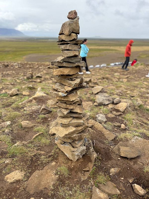 A tall stone cairn stands prominently in a rugged landscape with people in the background.