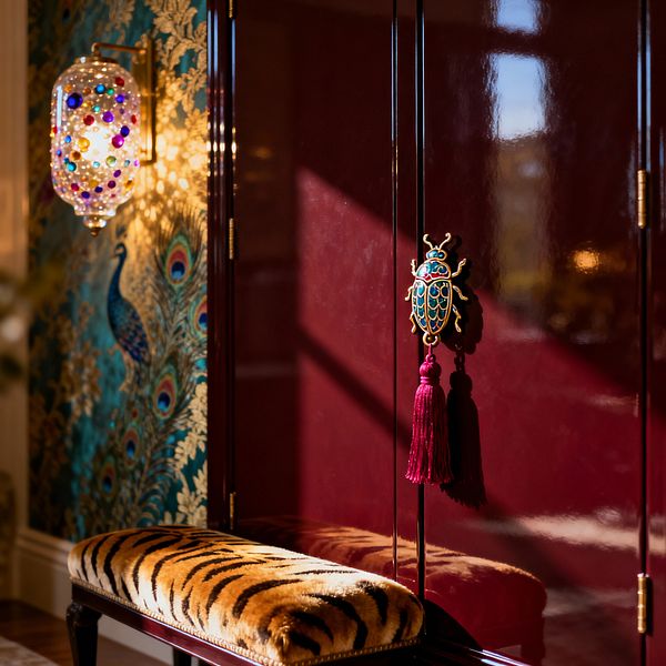 A jewel-toned bedroom corner features a high-gloss burgundy wardrobe with brass hinges and a blue-and-gold beetle pull finished with a magenta tassel. To the left, teal peacock wallpaper with metallic gold shines beside a brass sconce filled with multicolored glass orbs, while a dark-wood bench with plush tiger-print upholstery sits in front, all bathed in warm natural light.