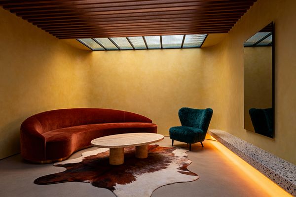 A contemporary living room with ochre plaster walls and a ribbed wood-slat ceiling features a linear skylight above a curved rust velvet sofa, an oval travertine coffee table with cylindrical legs, and a brown-and-white cowhide rug. A deep teal, fuzzy lounge chair sits beside a floating terrazzo bench with warm amber underlighting, while a large black-framed mirror and smooth concrete floor complete the minimalist, sculptural setting.