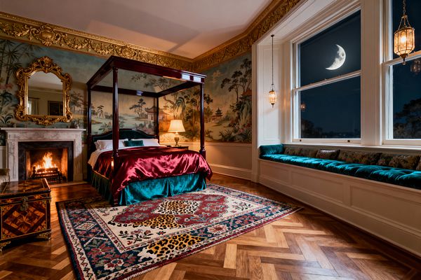 A richly layered bedroom centers on a mahogany four-poster bed dressed in burgundy satin and teal velvet, set against panoramic chinoiserie mural walls under an ornate gilded cornice. A marble fireplace with a gold-framed mirror glows at left, while a leopard-motif rug rests on herringbone wood flooring. To the right, a long teal velvet window seat spans large sash windows showing a crescent moon, with brass lantern pendants and a table lamp casting warm light.