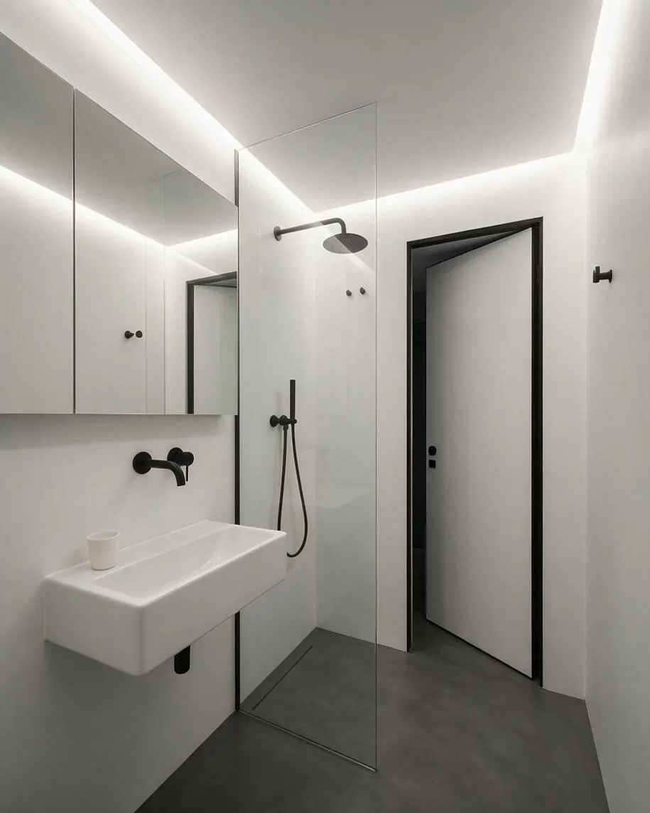 A compact minimalist master bathroom with white walls, a floating white sink, and a large mirrored cabinet on the left. A clear glass shower partition stands in the center, with a black rainfall showerhead and matte black fixtures contrasting against the pale surfaces and dark gray floor.