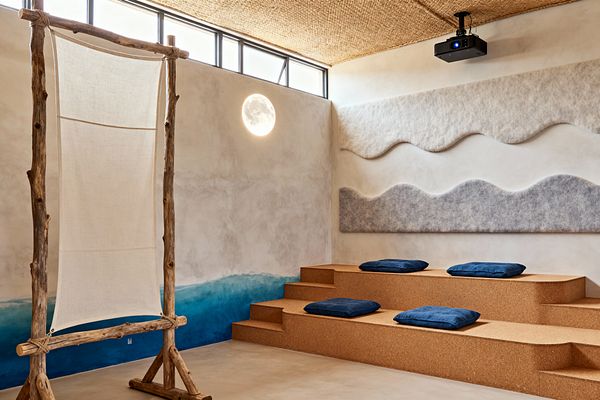 A serene media room with cork tiered seating topped by navy cushions faces a projector mounted to a woven natural-fiber ceiling. Textured plaster walls feature a blue ocean gradient, wave-shaped gray felt acoustic panels, and a round moon-like wall light, while a driftwood frame with a linen sail panel and slim clerestory windows reinforce the coastal, organic feel.