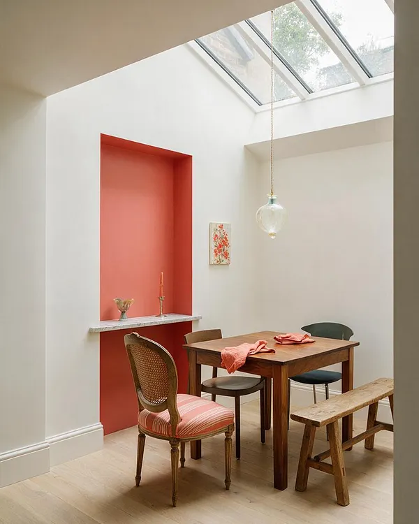 A bright eclectic dining nook with a small wooden table and mixed seating under a skylight. White walls, a coral recessed alcove with a marble ledge, and a clear glass pendant create a warm, airy atmosphere.
