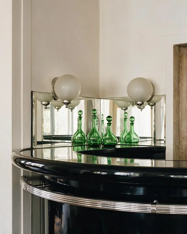 An Art Deco-inspired corner vignette features a glossy black lacquer vanity or console with a curved front and chrome ribbed trim. Above it, mirrored panels reflect a pair of frosted globe sconces and several small green glass decanters arranged in a neat cluster.