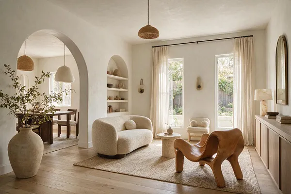A warm organic modern living room with rounded archways, a textured cream sofa, sculptural wooden lounge chair, and neutral rug. Sheer curtains soften tall windows while natural wood, plaster walls, woven lighting, and ceramic decor create a calm, sunlit atmosphere.