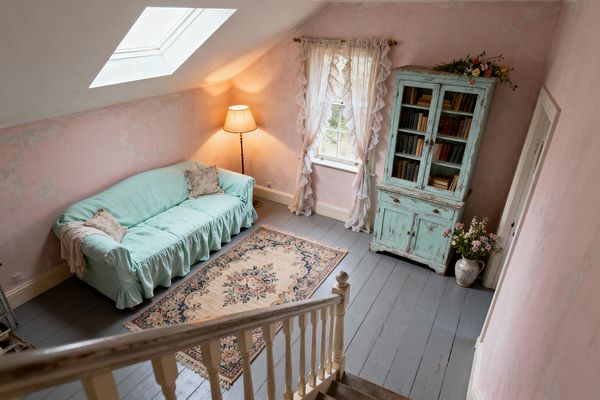 A cozy attic living room with a sloped ceiling and skylight features blush pink limewashed walls, a mint slipcovered sofa with a ruffled skirt, and a cream floral rug on gray-painted wood floorboards. A duck-egg blue distressed glass-front cabinet holds vintage books, topped with flowers, while a ceramic jug of blooms sits nearby. Sheer ruffled curtains and a brass floor lamp soften the light, with a worn white stair banister in the foreground.
