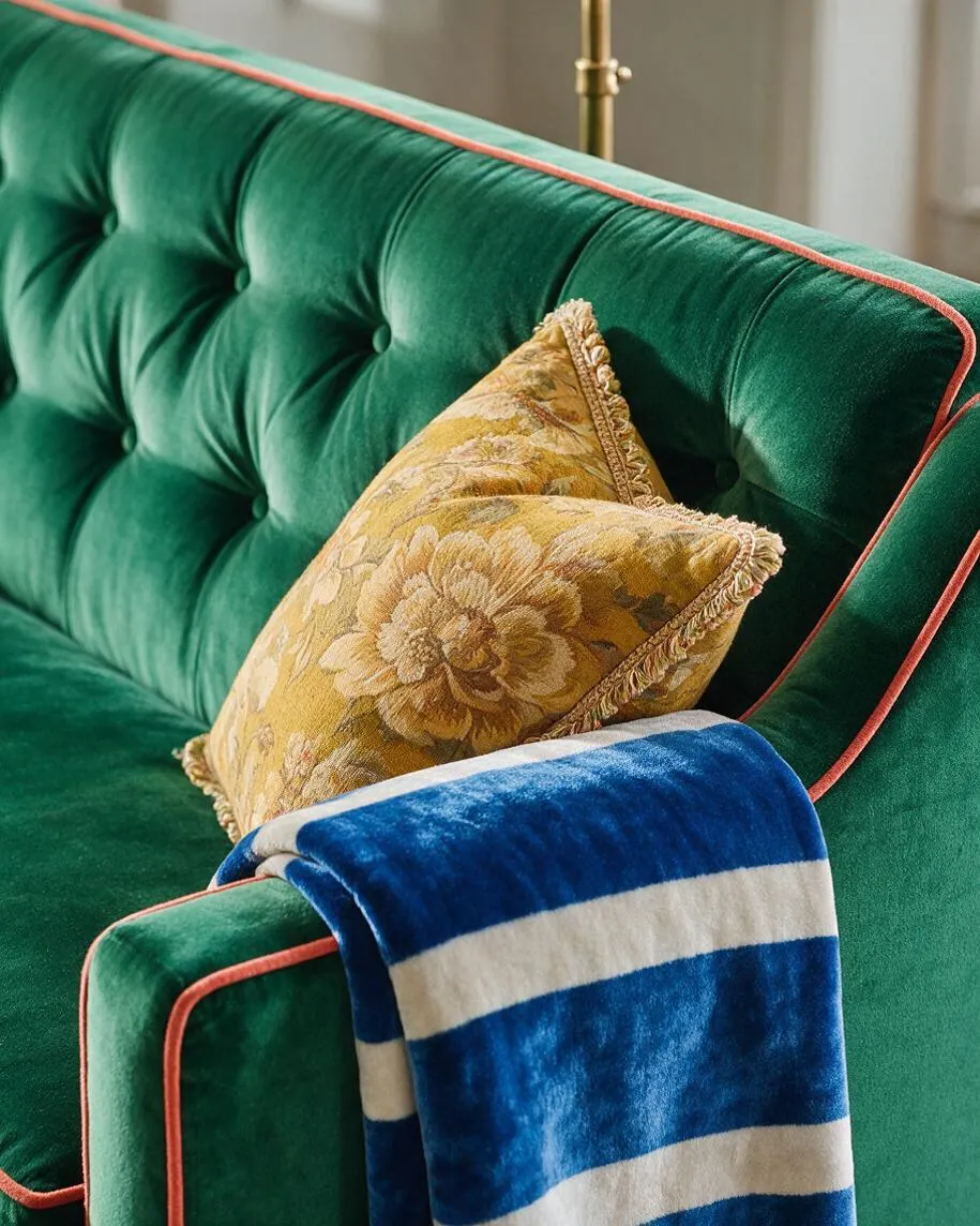 A close-up of a deep emerald green velvet sofa with tufted upholstery and coral piping along the edges. A gold floral cushion and a blue-and-white striped throw blanket add layered texture and rich color, creating a luxurious maximalist look.
