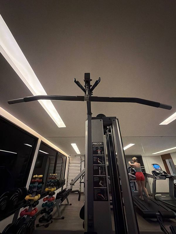 A woman in gym attire stands with a weightlifting bar, preparing for a workout in a gym setting.