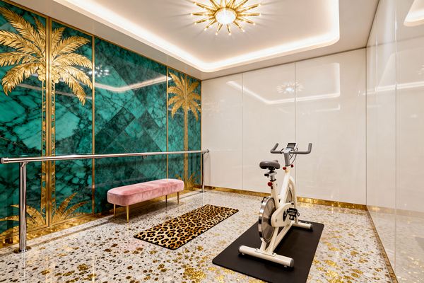 A Hollywood Regency home gym with an emerald malachite-pattern accent wall featuring gold palm motifs, a polished chrome ballet barre, and a blush velvet bench placed beside a leopard-print runner. The room has terrazzo flooring with gold flecks, glossy ivory lacquered wall panels, a brass sunburst chandelier set within cove lighting, gold baseboards, and a white stationary bicycle positioned on a black mat.